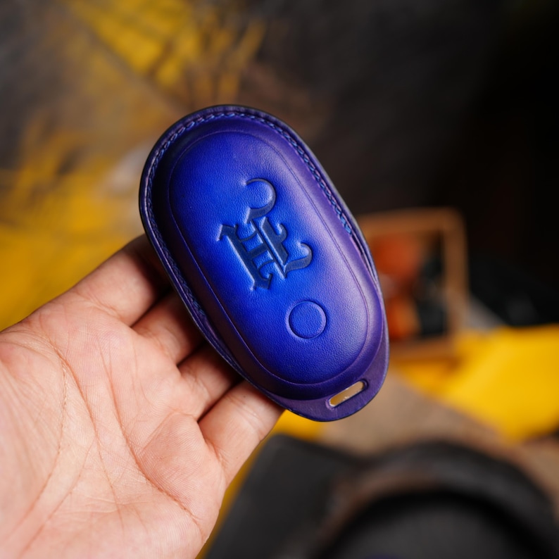 May include: A blue leather key fob cover with a debossed emblem. The cover is oval-shaped with a key ring hole and a circular indentation. The emblem is a stylized design. The leather has a gradient effect, transitioning from a darker to a lighter blue.
