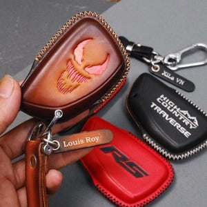 May include: A collection of leather key fob covers in various colors, including brown, red, and black. One brown cover features a carved design, while others display text. Keychains and tags with names are also included.