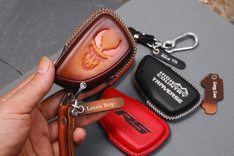 May include: A collection of leather key fob covers in various colors, including brown, red, and black. One brown cover features a carved design, while others display text. Keychains and tags with names are also included.
