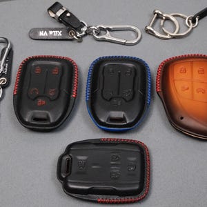 May include: An assortment of key fobs and keychains. Several black key fobs with red and blue stitching are displayed, along with a brown leather fob. Keychains include a silver carabiner and a black leather strap with the text "NA TIX".