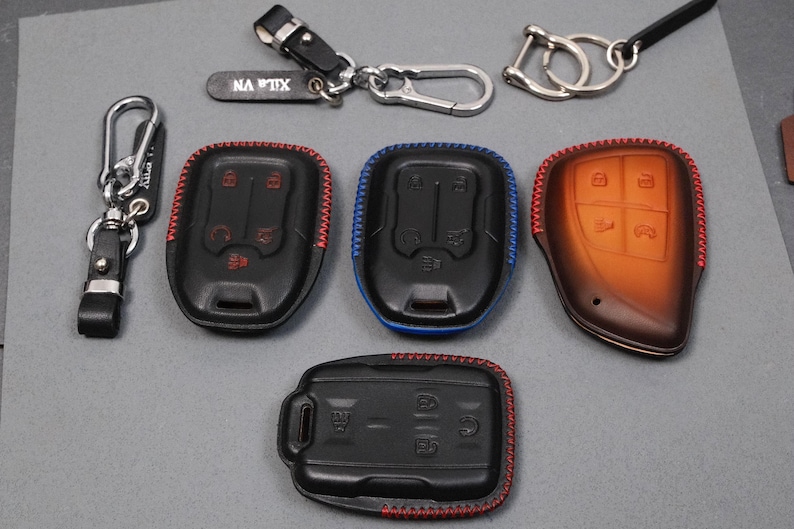 May include: An assortment of key fobs and keychains. Several black key fobs with red and blue stitching are displayed, along with a brown leather fob. Keychains include a silver carabiner and a black leather strap with the text "NA TIX".