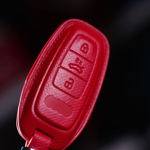 May include: A red leather car key fob cover. The cover has three buttons with embossed icons for locking, unlocking, and opening the trunk. The fob is a vibrant red color and has a textured surface. The background is black.