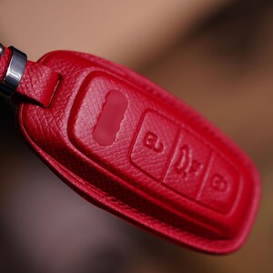 May include: A red leather car key fob cover with silver metal hardware. The fob has three embossed buttons and a key ring attachment. The leather has a textured appearance.