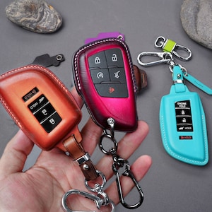 May include: Three leather key fobs with different colored stitching and button layouts. The key fobs are attached to metal key rings. The key fobs are on a hand.