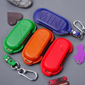 Può includere: Quattro portachiavi in ​​pelle con custodie di diversi colori per le chiavi dell'auto. Le custodie sono verde, arancione, rosso e blu. La custodia blu ha la parola "MINI" in rilievo. I portachiavi sono su una superficie grigia.