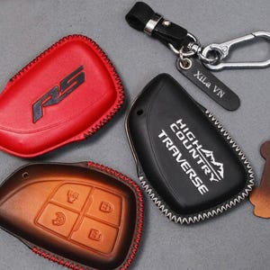 May include: A collection of leather key fob covers in red, brown, and black. The red cover has a black RS logo, the black cover has "HIGH COUNTRY TRAVERSE" text, and the brown cover has key symbols. A keychain with a tag that says "Xila VN" is also included.