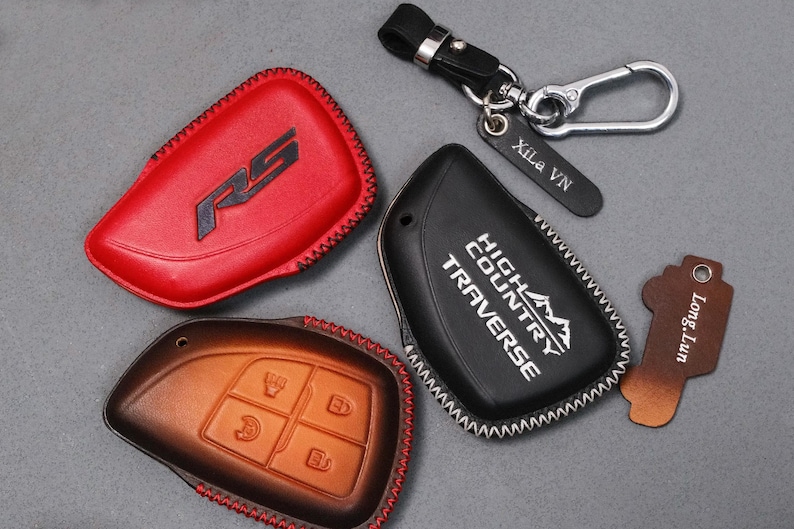 May include: A collection of leather key fob covers in red, brown, and black. The red cover has a black RS logo, the black cover has "HIGH COUNTRY TRAVERSE" text, and the brown cover has key symbols. A keychain with a tag that says "Xila VN" is also included.