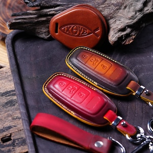 May include: Leather key fobs in shades of brown and red, with yellow stitching and silver key rings. One fob features an Ichthys symbol. The key fobs are on a dark wooden tray.