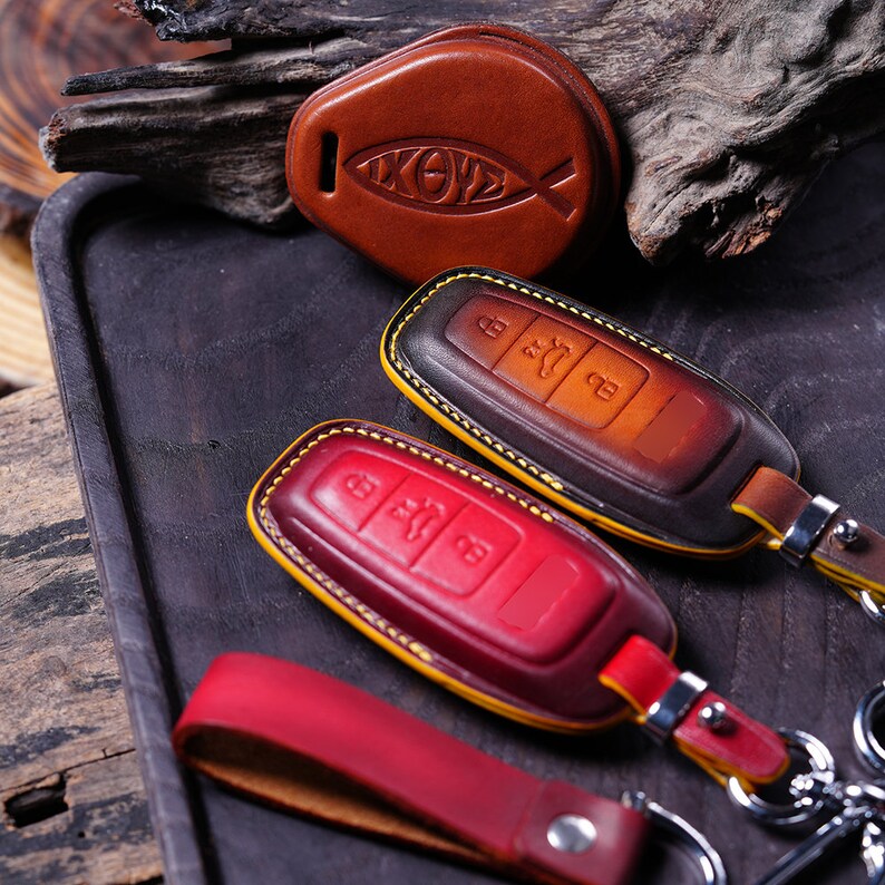 May include: Leather key fobs in shades of brown and red, with yellow stitching and silver key rings. One fob features an Ichthys symbol. The key fobs are on a dark wooden tray.