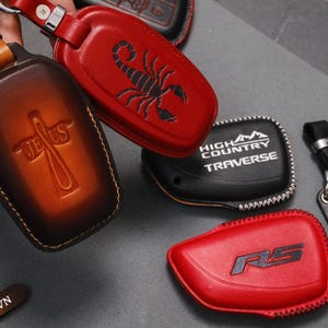 May include: A collection of leather key fob covers in various colors. One is brown with a cross and the word "Jesus", another is red with a scorpion, and a third is black with "High Country Traverse". Other key fobs are red with "RS" and black with "Xila VN".