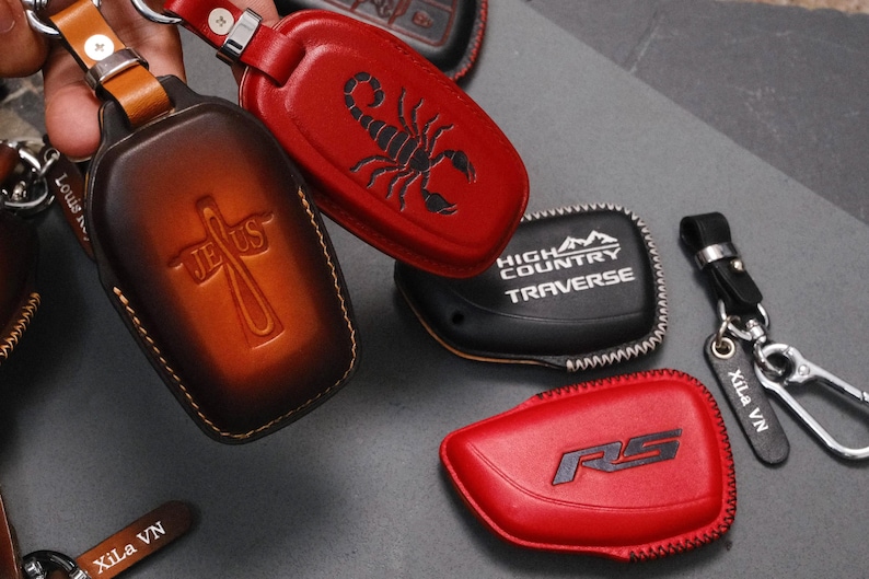 May include: A collection of leather key fob covers in various colors. One is brown with a cross and the word "Jesus", another is red with a scorpion, and a third is black with "High Country Traverse". Other key fobs are red with "RS" and black with "Xila VN".