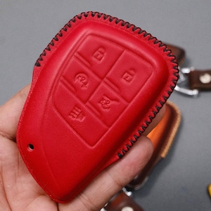 May include: A bright red leather key fob cover with black stitching. The cover has embossed icons for lock, unlock, and other functions. The key fob cover is a protective accessory for car keys.