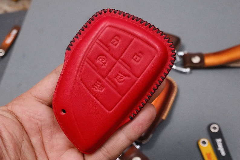May include: A bright red leather key fob cover with black stitching. The cover has embossed icons for lock, unlock, and other functions. The key fob cover is a protective accessory for car keys.