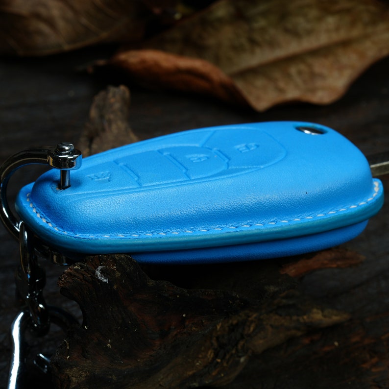 Puede incluir: Un llavero de cuero azul con un anillo y una cadena de metal plateado. El llavero tiene un borde cosido y est&aacute; dise&ntilde;ado para proteger una llave de coche.