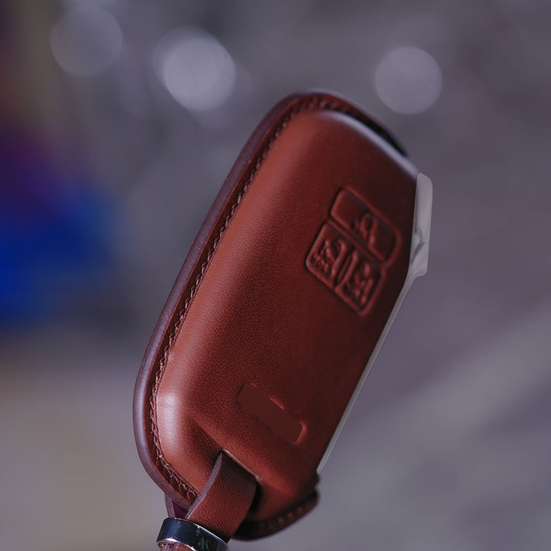 May include: A brown leather key fob with a stitched edge and a small rectangular button. The fob has a loop for attaching to a keychain.