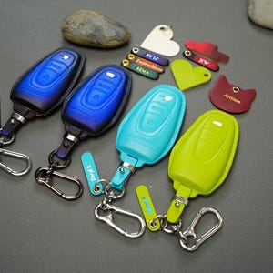 May include: Four keychains with different colored leather key covers. The key covers are blue, green, turquoise, and purple. Each keychain has a metal ring and a small metal tag with text on it.
