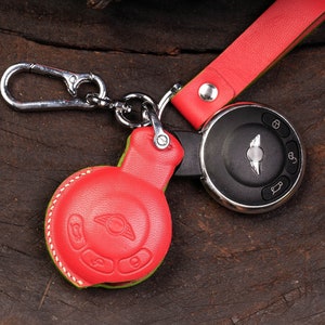 Puede incluir: Un llavero de cuero rojo con una cadena de plata y una llave negra con el logotipo de Mini Cooper. El llavero tiene una costura verde alrededor del borde.