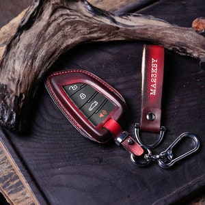 May include: A red leather key fob with a silver key ring and a brown leather strap with the name "MA23KSY" embossed on it.