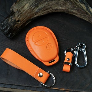 May include: Orange leather key fob with two buttons and a silver metal loop. The fob is on a wooden surface with a piece of wood in the background.