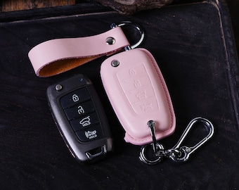 May include: A pink leather key fob with a silver key ring and a black car key with multiple buttons. The key fob has a silver button and the key has the words "HOLD" and "UNLOCK" printed on it.