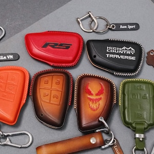 May include: A collection of leather key fob covers in various colors, including red, orange, brown, black, and green. Some covers feature embossed designs and text, such as "RS", "HIGH COUNTRY TRAVERSE", and a skull. Keychains and key rings are also included.