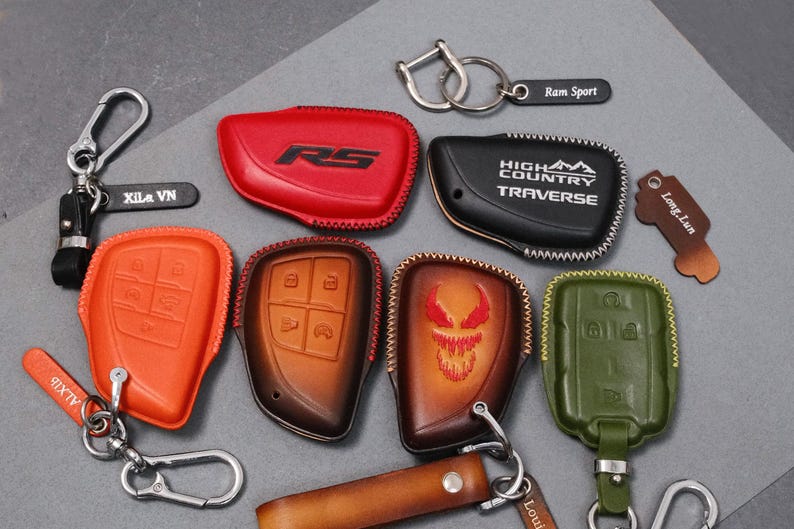 May include: A collection of leather key fob covers in various colors, including red, orange, brown, black, and green. Some covers feature embossed designs and text, such as "RS", "HIGH COUNTRY TRAVERSE", and a skull. Keychains and key rings are also included.