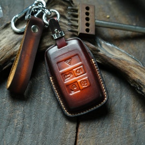May include: A brown leather key fob with stitching around the edges. The fob has a key chain attached and features a four-button design with symbols for unlock, lock, boot, and panic.