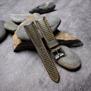Könnte beinhalten: Zwei grüne Lederarmbänder mit orangefarbenen Nähten. Die Armbänder liegen auf einer grauen, strukturierten Oberfläche mit mehreren grauen Steinen.