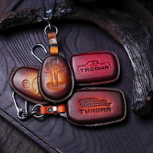 May include: A collection of handcrafted leather keychains and key fobs in shades of brown and red. One keychain features a cross design, while others display the "TACOMA" and "TUNDRA" logos. The key fobs have metal clasps.