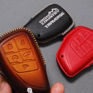 May include: A collection of leather key fob covers in various colors. One is brown with embossed key symbols, another is black with "HIGH COUNTRY TRAVERSE" text, and a third is red. All have decorative stitching.