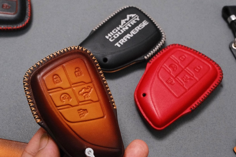 May include: A collection of leather key fob covers in various colors. One is brown with embossed key symbols, another is black with "HIGH COUNTRY TRAVERSE" text, and a third is red. All have decorative stitching.