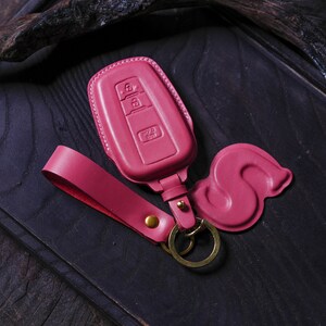 May include: A pink leather key fob with a key ring and a decorative pink leather charm. The key fob has a rectangular shape and features three buttons.