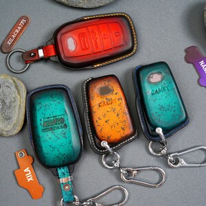 May include: Four leather keychains with different colored designs. The keychains feature the words "CAMRY" and "GR GARAGE" on them. The keychains are attached to silver key rings.