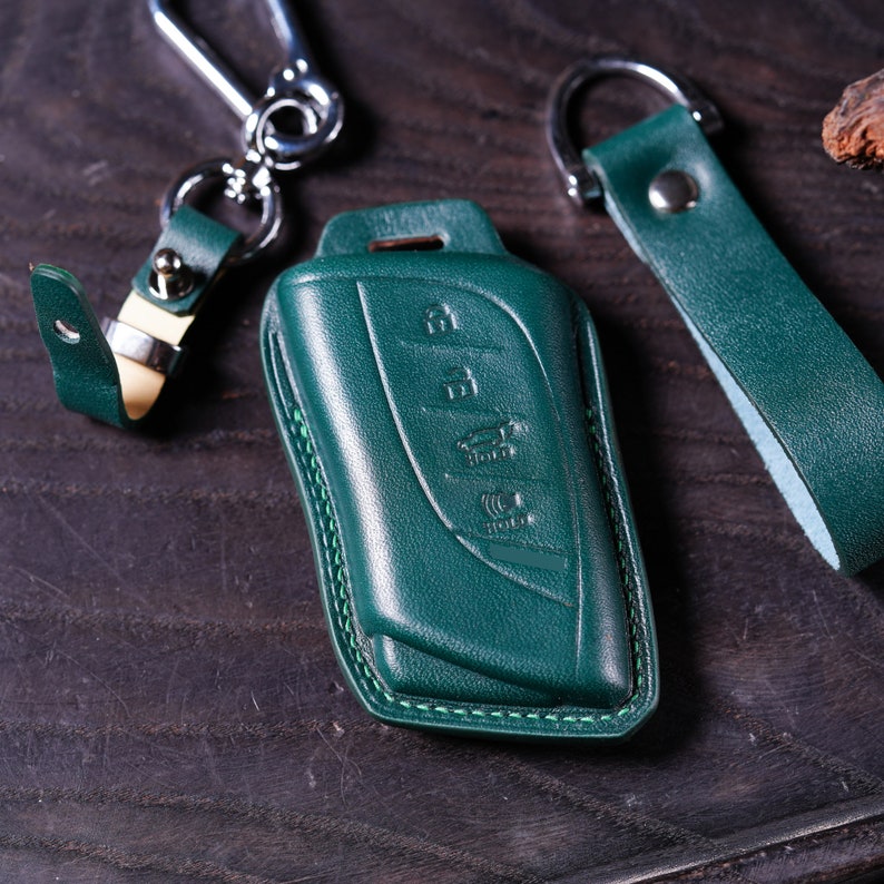 May include: A dark green leather key fob with stitching around the edges. The fob has a design of four buttons on the front.