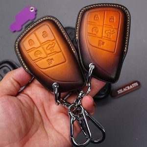 May include: A brown leather key fob with a silver keychain and a silver clasp. The key fob has a stitched design and is embossed with buttons. The keychain has a small tag that says "Xilacrafts".
