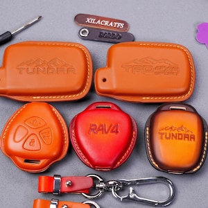 Könnte beinhalten: Eine Sammlung von Leder-Schlüsselanhänger-Hüllen in verschiedenen Formen und Farben. Die Hüllen sind mit den Wörtern "TUNDRA", "RAV4" und "TRD 4x4" geprägt. Ein Schlüsselanhänger und ein kleiner Schraubendreher sind ebenfalls sichtbar.