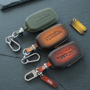 May include: Three leather key fob covers with metal clips. Two are brown gradient with Tundra and Tacoma TRD Off Road branding. One is olive green with Tundra branding. Two small leather tags are included, one labeled J.L.S DB12 Ullo and one Billie.