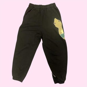 Op de afbeelding: Zwarte joggingbroek met elastische tailleband en manchetten. Een grote, kleurrijke slangengrafiek staat op het linkerbeen. De slang heeft een bruin lichaam, een groen en blauw vlak en een gele ster.