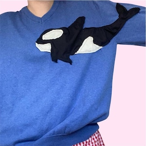 Op de afbeelding: Een blauwe V-hals sweatshirt met een geappliqueerd orka walvis ontwerp. De walvis is zwart en wit, met een gedetailleerde omtrek. De sweatshirt is effen, middelblauw van kleur en de walvis is gepositioneerd op de linkerbovenborst.