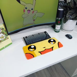 May include: A yellow Pikachu-themed desk mat with black eyes, red cheeks, and an orange mouth. A black and grey keyboard sits above the mat. A computer monitor displays a Pikachu graphic. A bottle of gin and a mouse are also on the desk.