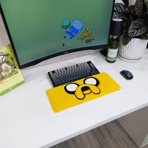 May include: A yellow Jake the Dog mousepad with black and white details, placed on a white desk. A black and gray keyboard sits above the mousepad. A computer monitor displays cartoon characters. A black mouse and a calendar are also visible.