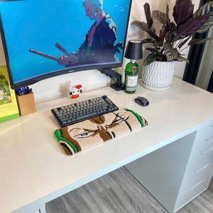 Pode incluir: Uma mesa branca com um monitor de computador exibindo arte de anime. Um teclado e um tapete com o rosto de um personagem estão na mesa. Uma garrafa verde, uma lâmpada preta e uma planta em vaso também são visíveis.