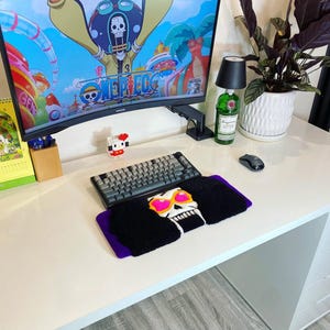 May include: A black and purple keyboard wrist rest featuring a skull design with pink sunglasses and black hair. A gray keyboard sits above the wrist rest. A computer monitor displays a colorful cartoon image.