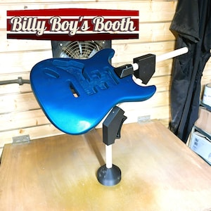 May include: A blue electric guitar body is mounted on a rotating stand, ready for painting. The stand has a white pole and a black base. The guitar body is in a spray booth, with a black drop cloth in the background.