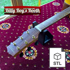 Op de afbeelding: Close-up van de kop van een akoestische gitaar met een zwarte gitaarstandaard. De gitaar is een Turner Guitars model. De kop is gemaakt van hout en heeft een natuurlijke afwerking. De gitaar ligt op een rood tapijt met een bloemenpatroon. De tekst "Billy Boy's Booth" is zichtbaar op de achtergrond.