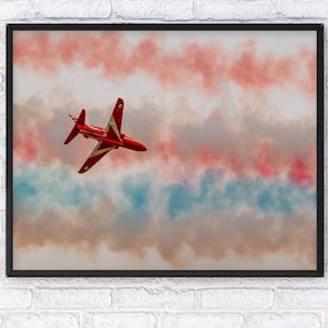 May include: A framed print featuring a red jet aircraft against a backdrop of red, white, and blue smoke trails. The jet has white accents and is angled in flight, creating a dynamic composition. The artwork is displayed in a black frame.
