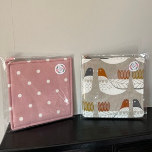Puede incluir: Dos agarraderas cuadradas en embalaje transparente. Una es rosa con lunares blancos. La otra es gris con un estampado de pájaros en blanco, naranja y negro. Ambas tienen una pequeña pegatina redonda.