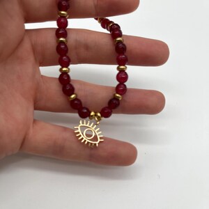 May include: A beaded bracelet featuring deep red, round beads and gold-colored spacers. A gold-colored charm in the shape of an eye hangs from the bracelet. The bracelet is held against a white background.