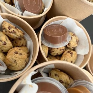 May include: Multiple round tan paper cups filled with small chocolate chip cookies and a clear plastic container with a brown lid labelled Darnell. The cookies are golden brown with visible dark chocolate chips, nestled on white paper liners.