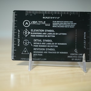 May include: Black metal ruler with a scale of 1/4 inch to 10 inches, 1/2 inch to 10 inches, and 3/4 inch to 1 inch. The ruler has symbols for view title, elevation, detail, and revision. The ruler also has a centre line that indicates the centre of an object. The ruler is made by www.cdgikhart.com.